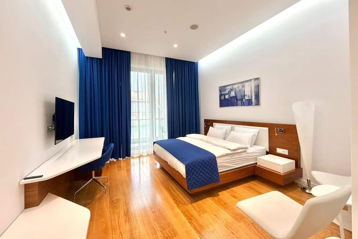 A modern hotel room with a wooden floor, a bed with a blue blanket, a desk with a chair, a bedside table, and a TV mounted on the wall.