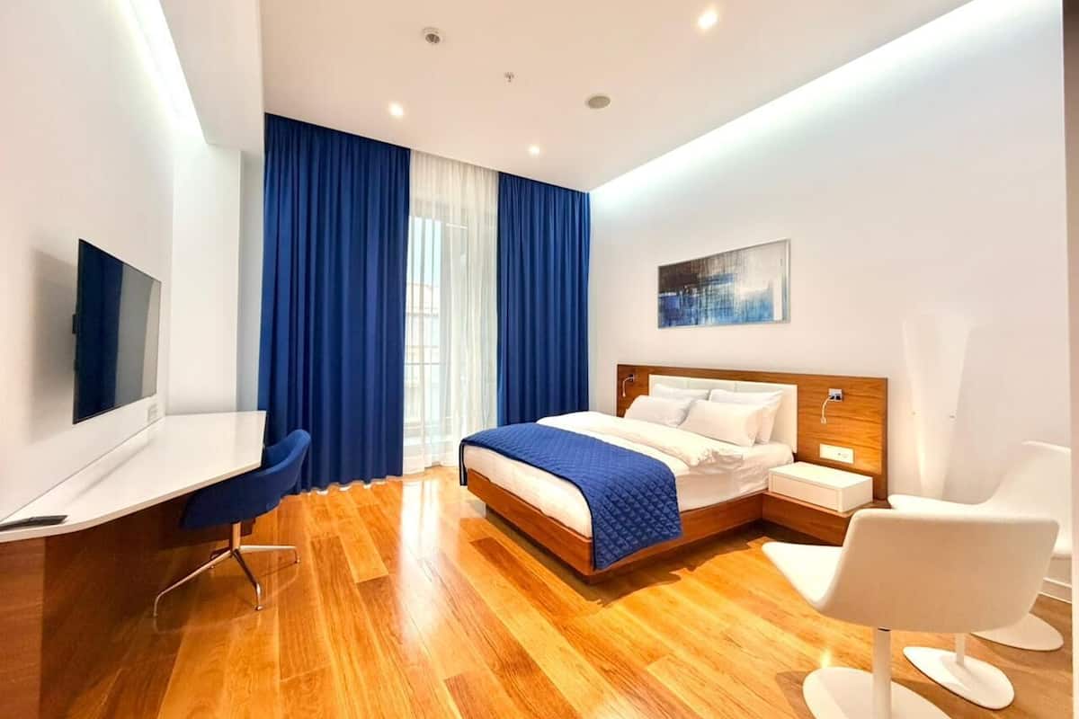A modern hotel room with a wooden floor, a bed with white bedding, a desk with a chair, and a TV on the wall.
