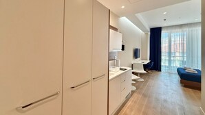 A modern hotel room with a large wardrobe, a small kitchenette, a seating area with a sofa and chairs, and a large window with curtains. - Condo Studio Plaza (Budva)