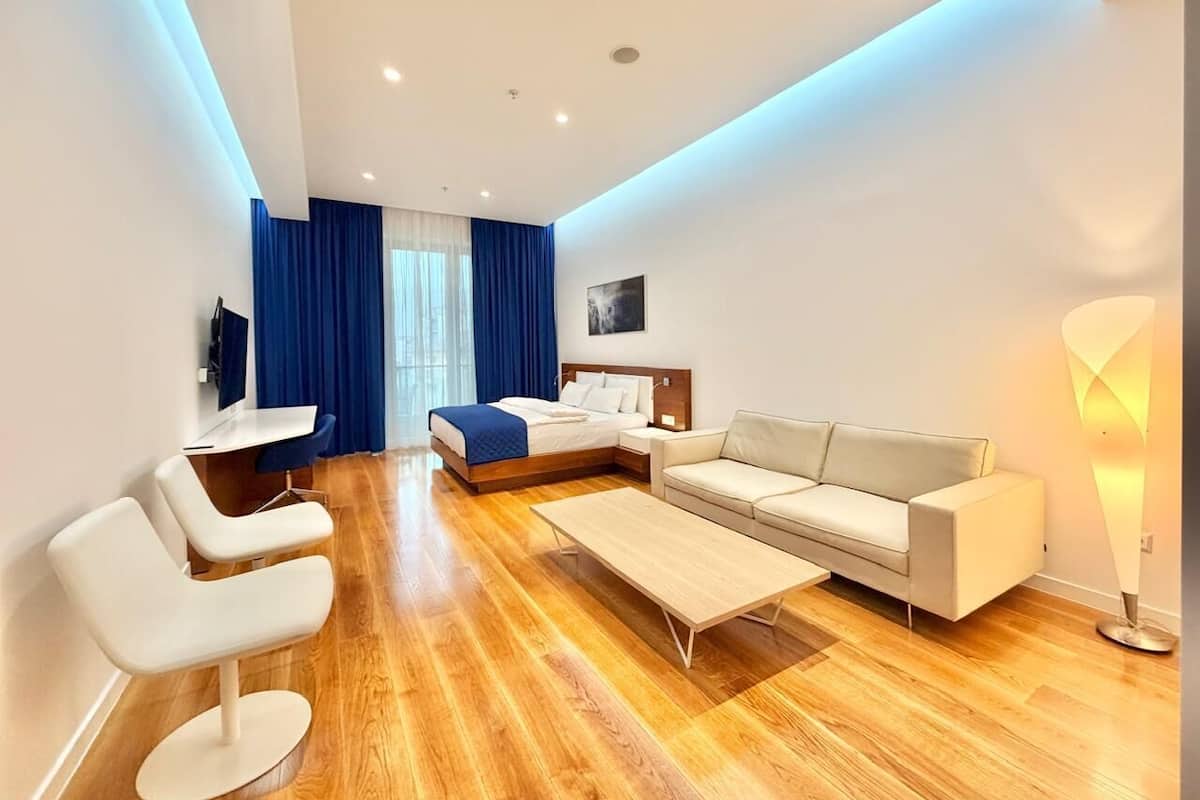 A modern hotel room with a wooden floor, a bed with a blue headboard, a white sofa, a small table, and a desk with a chair.