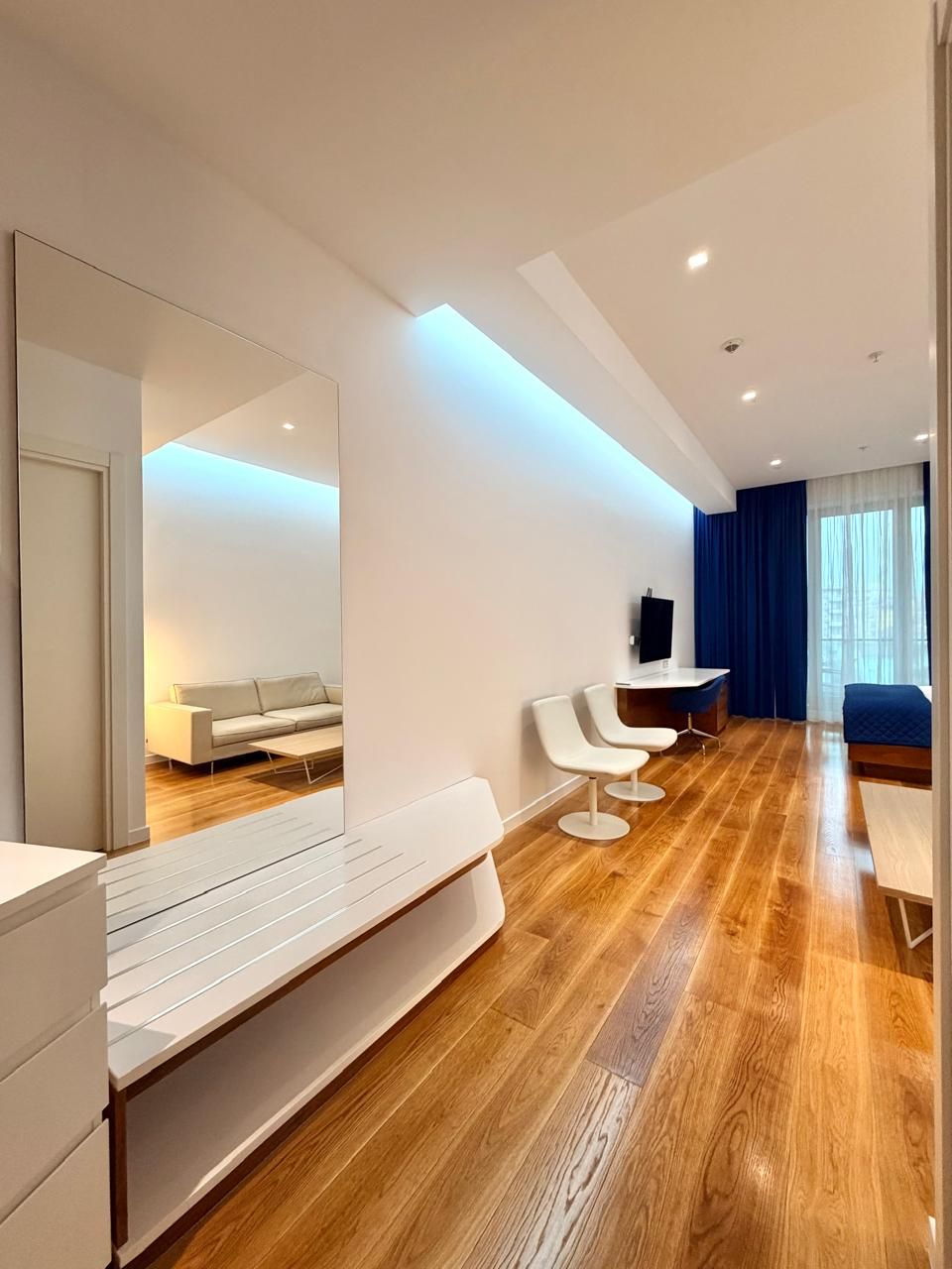 A modern hotel room with a wooden floor, a large mirror, a sofa, a desk, and a TV.
