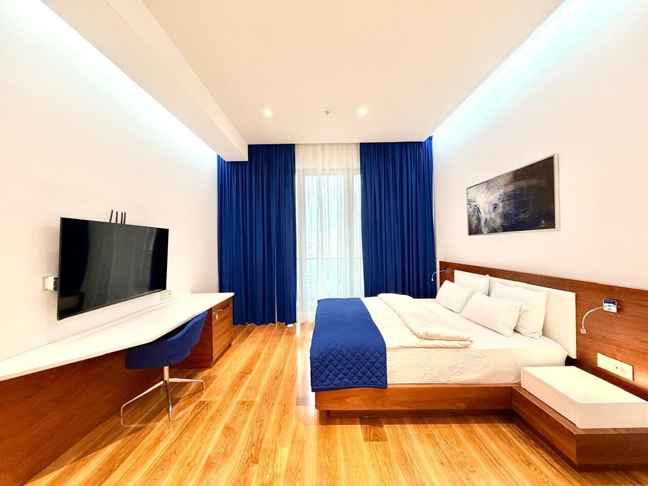 A modern hotel room with a wooden floor, a bed with white bedding, a desk with a flat-screen TV, and a large window with blue curtains.
