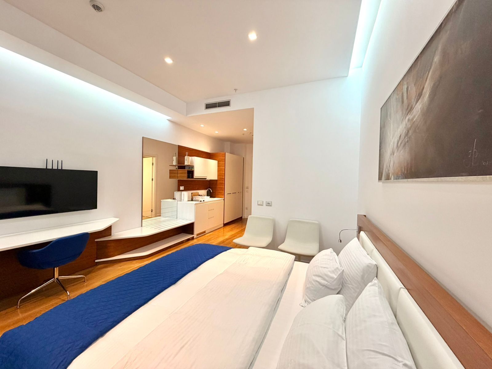 A modern hotel room with a large bed, a desk, a flat-screen TV, a kitchenette, and a painting on the wall.