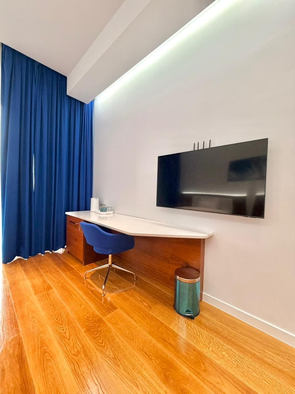 A modern room with a wooden floor, a white desk with a blue chair, a mounted TV, and a blue curtain.