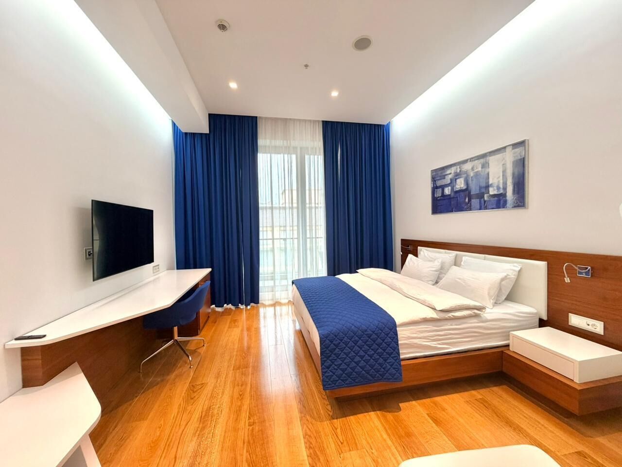 Una habitación de hotel moderna con piso de madera, una cama con manta acolchada, un escritorio con televisión de pantalla plana y un ventanal con cortinas azules.