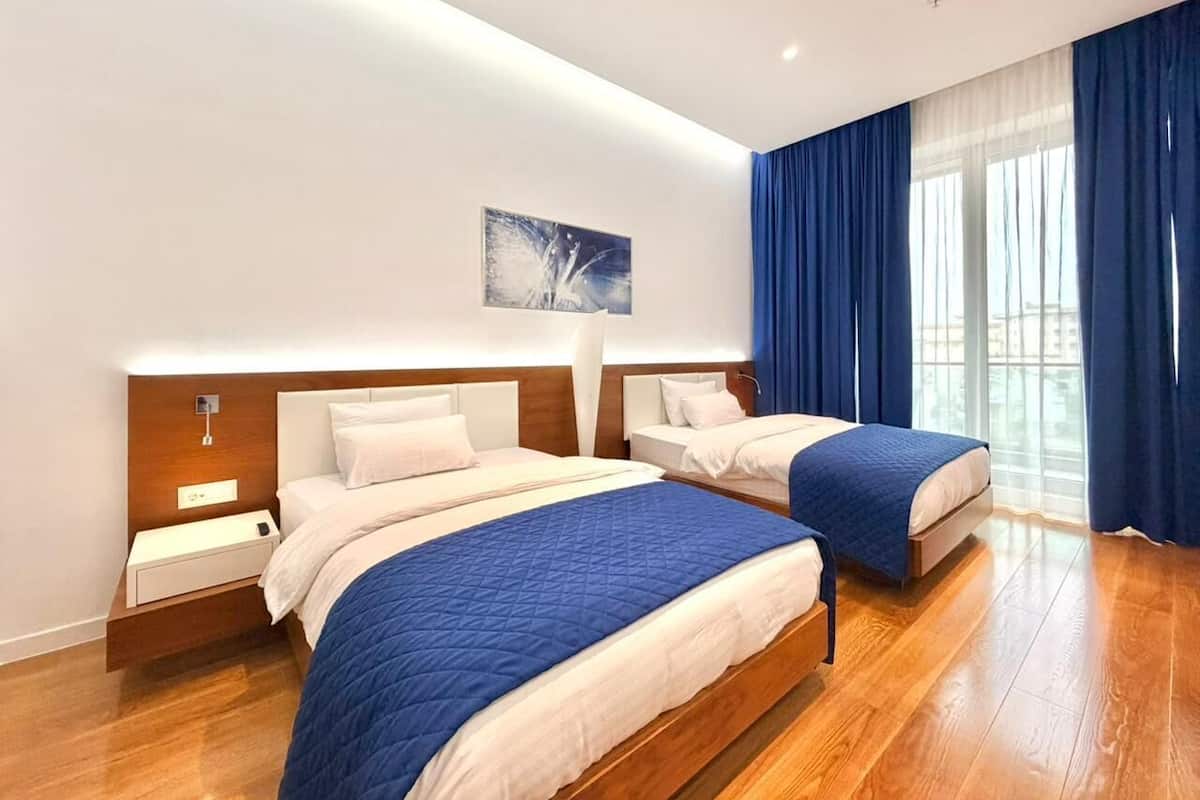 A modern hotel room with two beds, wooden flooring, and a large window with blue curtains.