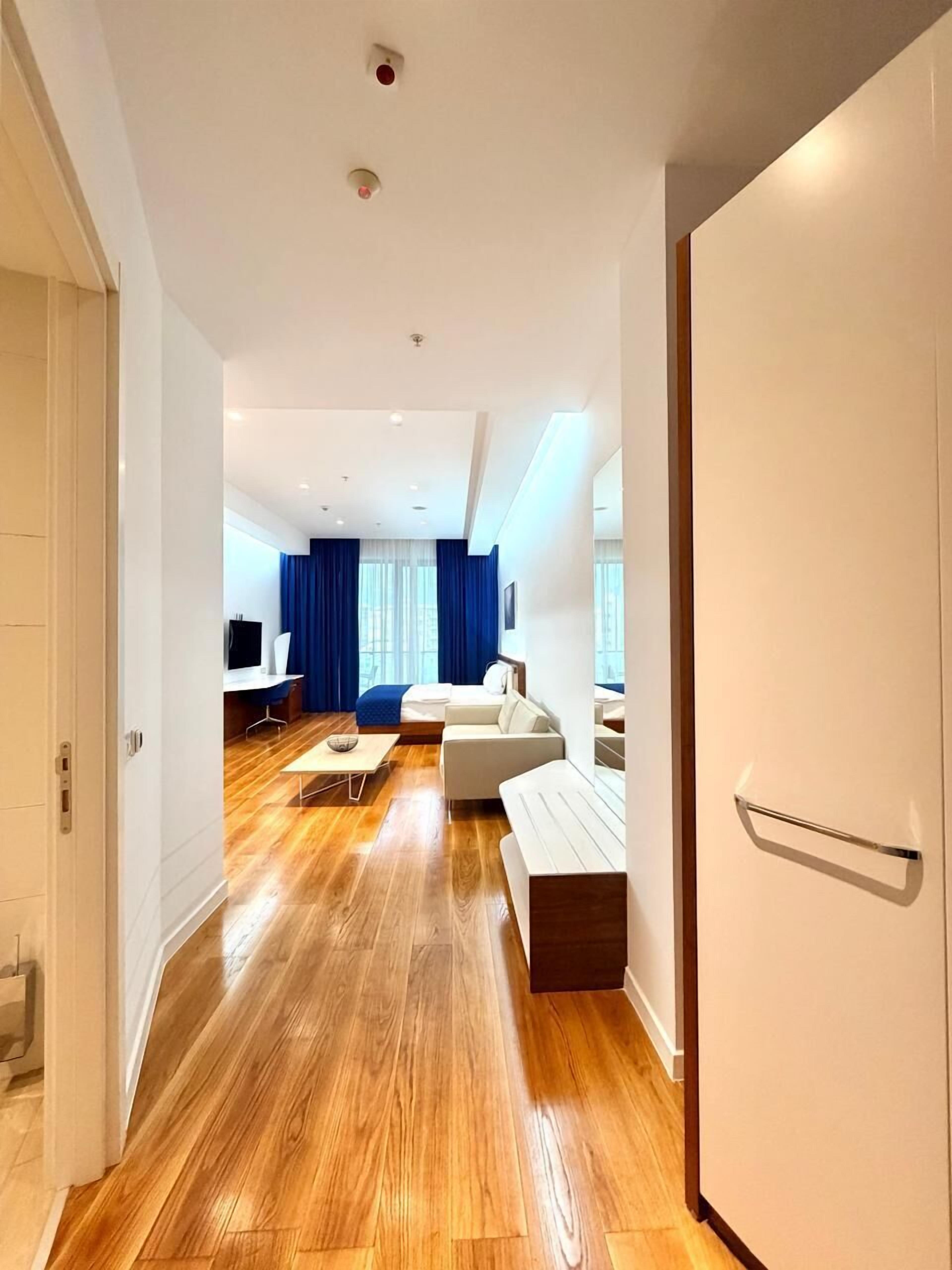 A modern hotel room with a wooden floor, a sofa, a TV, and a large window with blue curtains.