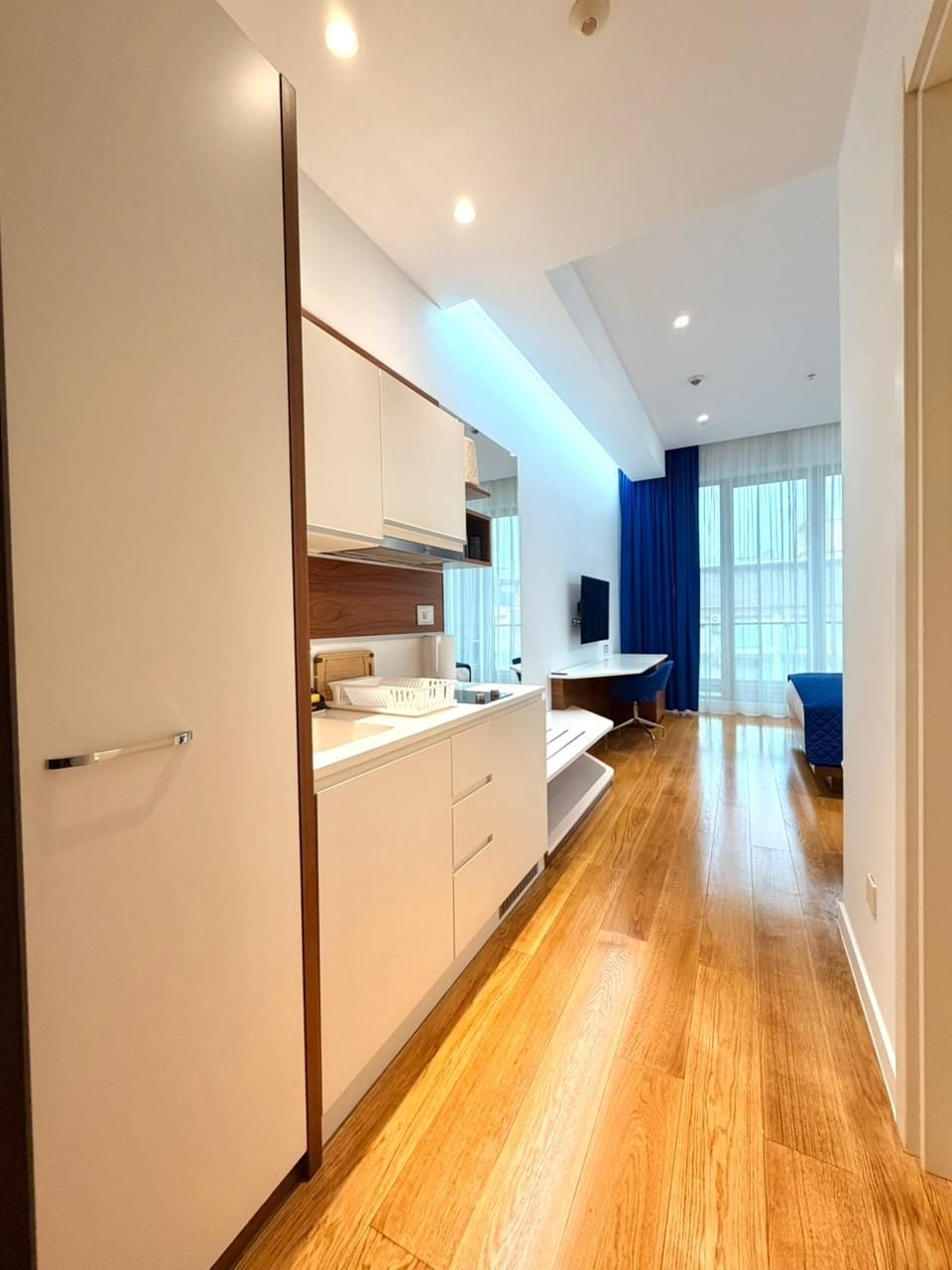 Una habitación de hotel moderna con kitchenette, zona de comedor y un ventanal con cortinas.