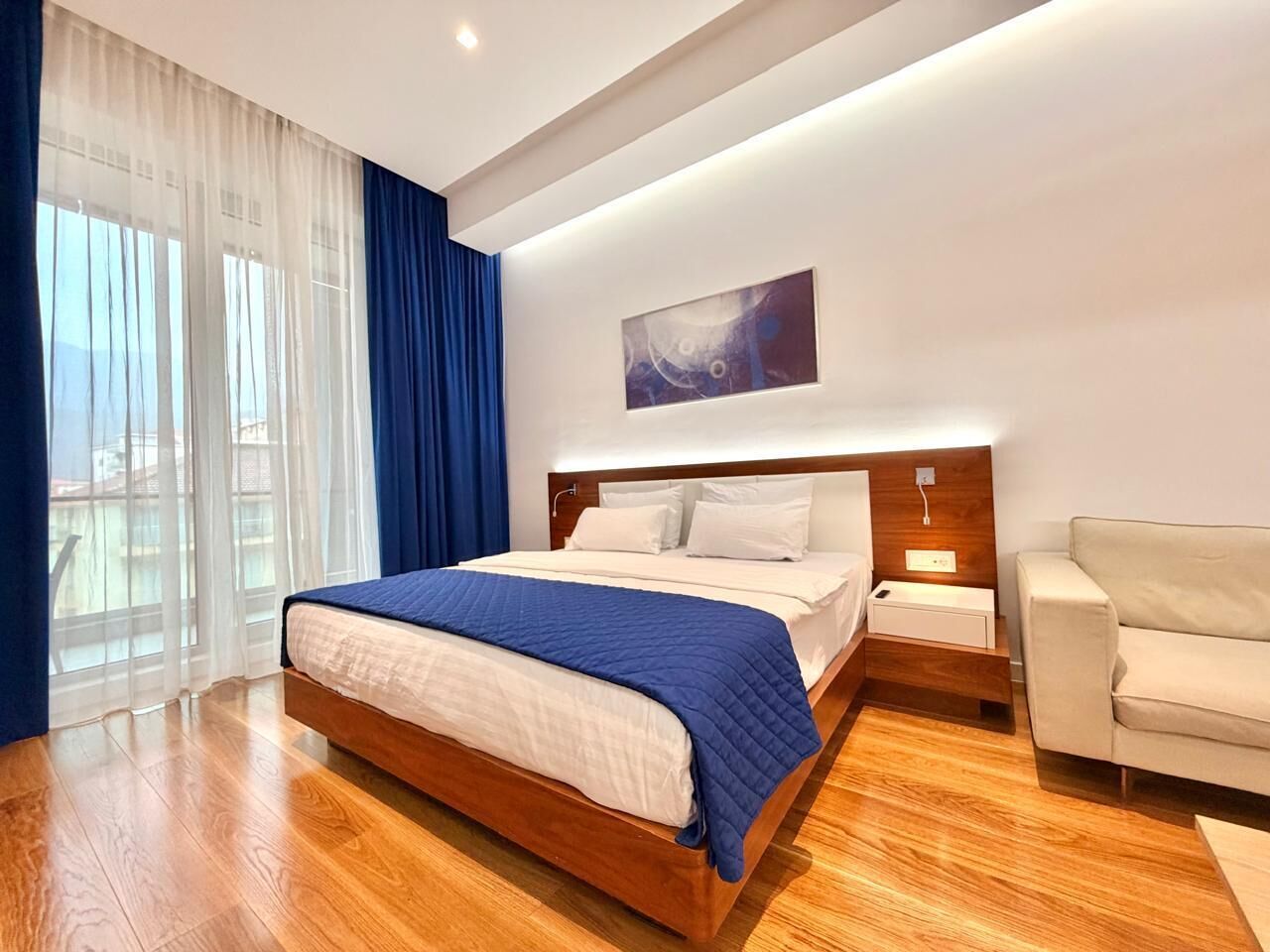 Une chambre d’hôtel moderne avec un grand lit, un plancher en bois, une table de chevet, un canapé et une grande fenêtre avec des rideaux.