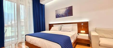 Une chambre d’hôtel moderne avec un grand lit, un plancher en bois, une table de chevet, un canapé et une grande fenêtre avec des rideaux.