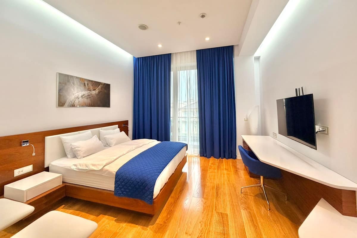 A modern hotel room with a bed, a TV, a desk, and a window with blue curtains.