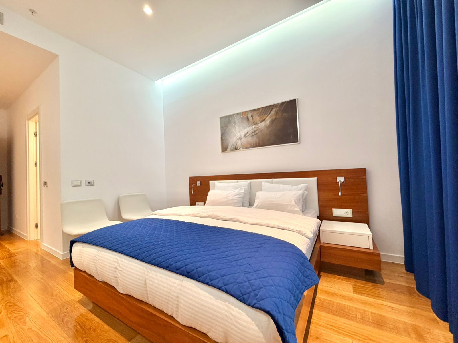 Una habitación de hotel moderna con cama de madera, ropa de cama blanca, un detalle en azul, una mesita de noche, una silla y un cuadro en la pared.