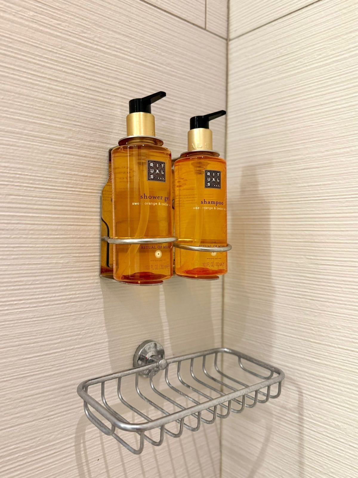 Zwei Flaschen Duschgel und Shampoo von Rituals auf einem an der Wand befestigten Metallregal in einem Badezimmer.