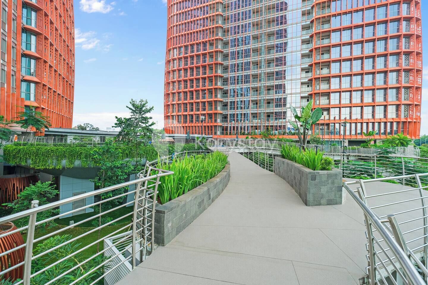 A modern urban walkway with greenery, metal railings, and high-rise buildings in the background.