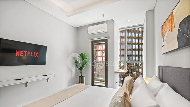A modern bedroom with a large bed, a TV mounted on the wall, a potted plant, and a view of the city through a window. - Taveli by Kozystay - TB Simatupang (Jakarta)
