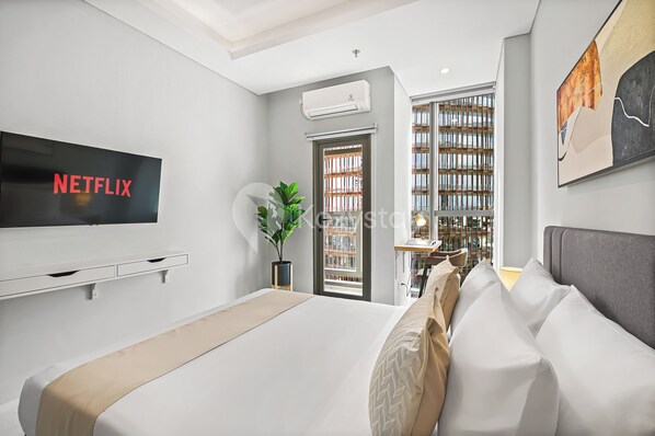 A modern bedroom with a large bed, a TV mounted on the wall, a potted plant, and a view of the city through a window. - Taveli by Kozystay - TB Simatupang (Jakarta)