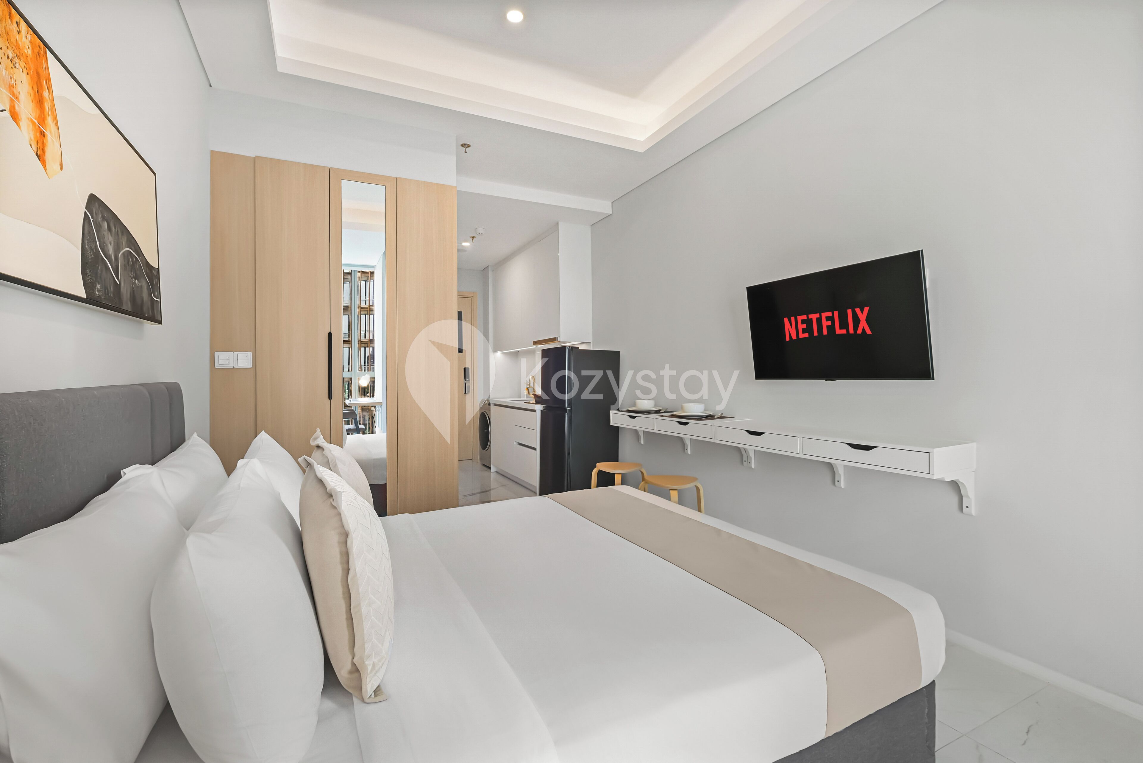 A modern hotel room with a bed, a desk, a TV displaying 'NETFLIX', and a view into the kitchen area.