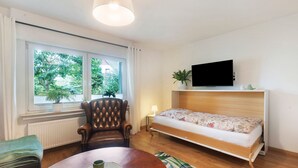 Ein Zimmer mit Bett, Fernseher, Sessel, Tisch und einem Fenster mit Blick ins Grüne.