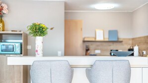 Moderni keittiö, jossa on valkoinen työtaso, kaksi harmaata baarituolia ja maljakko, jossa on keltaisia kukkia.