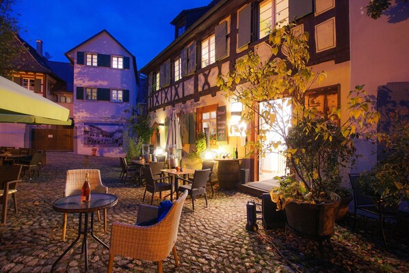 A traditional European courtyard with stone flooring, outdoor seating, and a building with exposed timber framing.