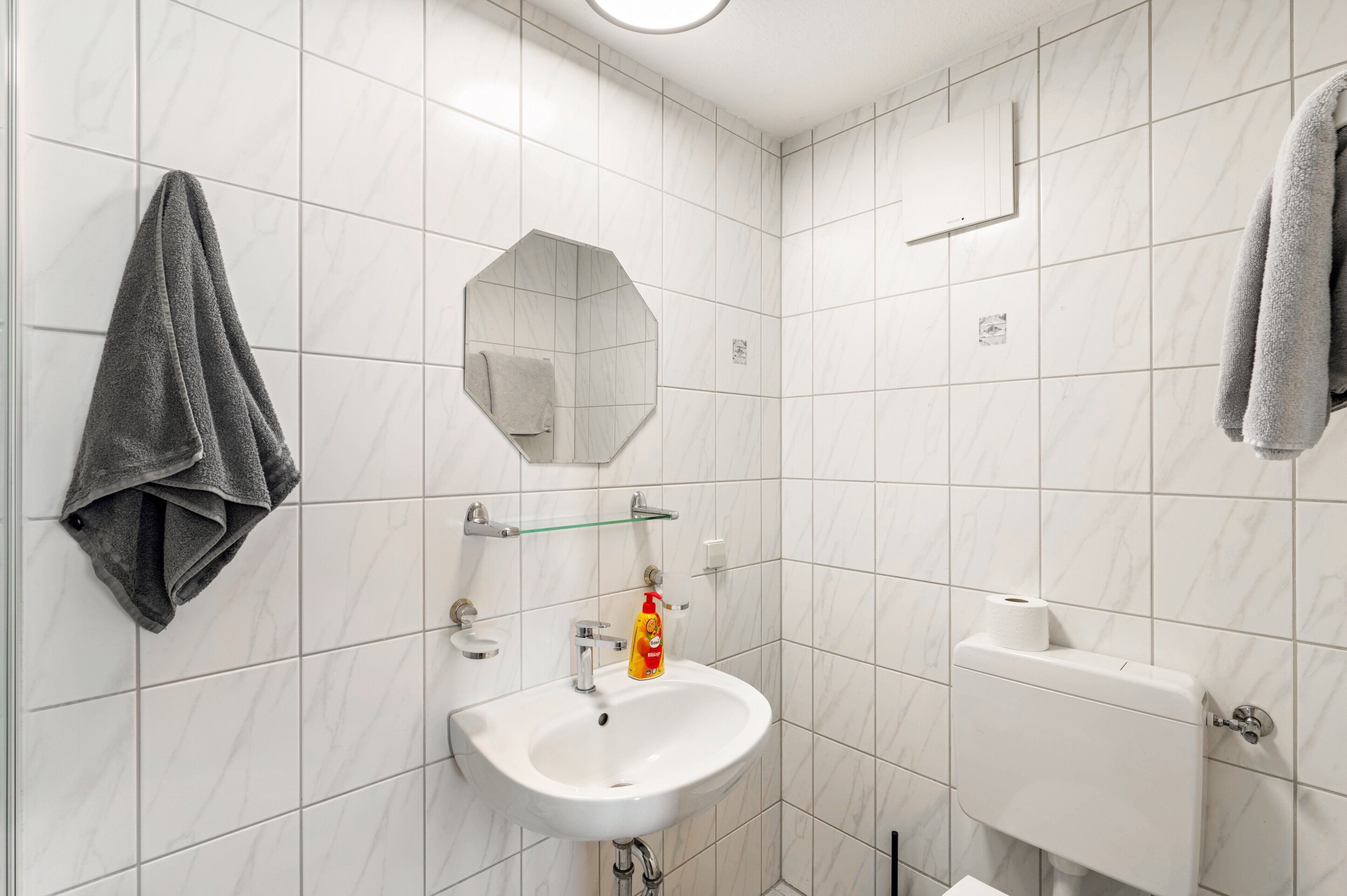 Un baño con baldosas de mármol blanco, un lavamanos blanco, un espejo, un perchero para toallas y un dispensador de papel higiénico.