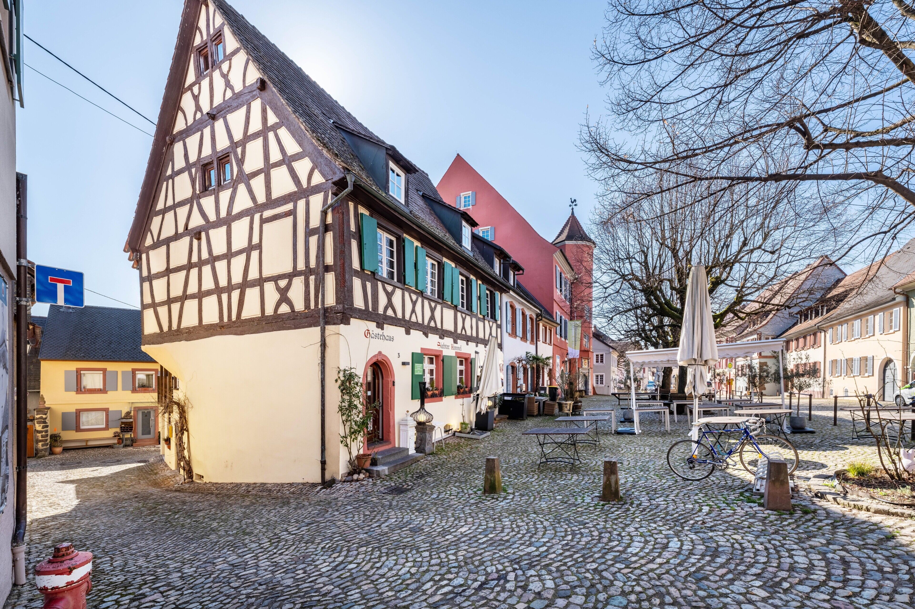 A cobblestone street with traditional half-timbered buildings, a covered seating area, and a bicycle.