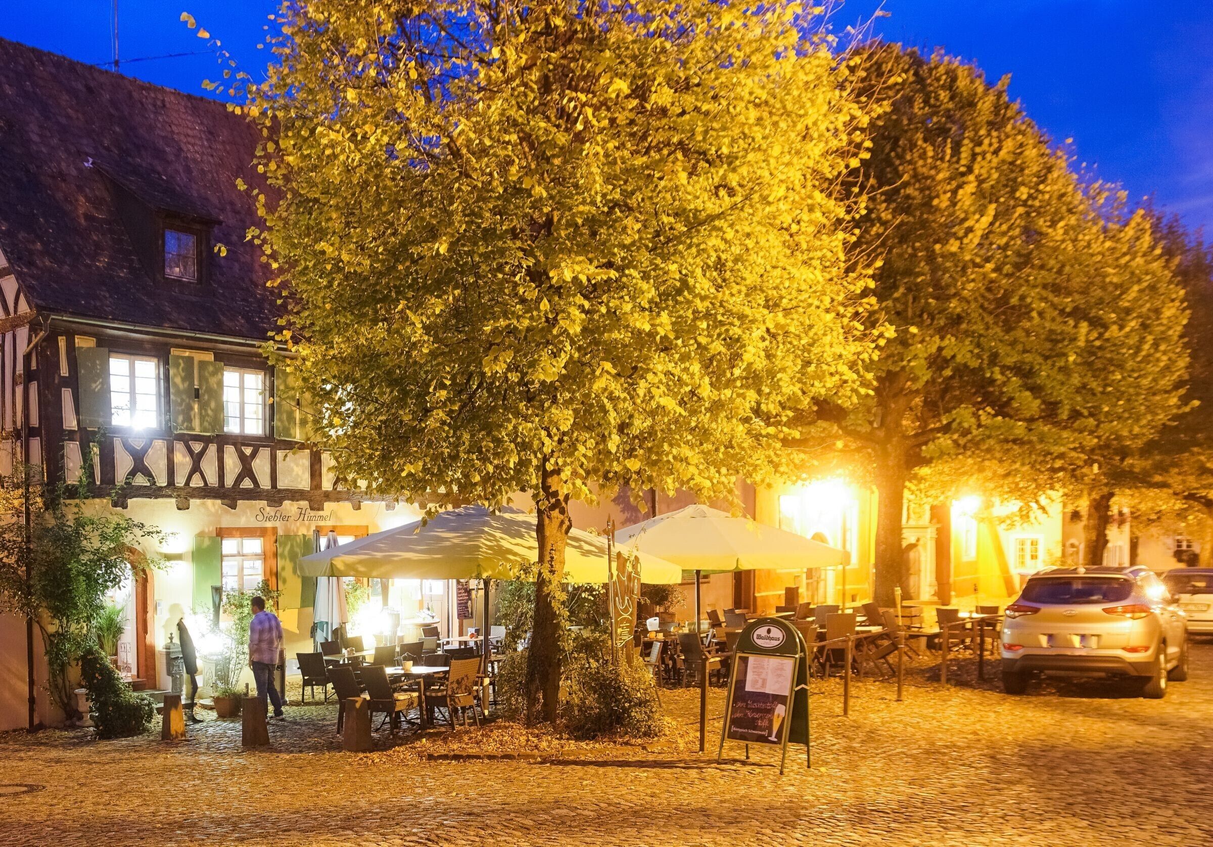 Una calle adoquinada con mesas al aire libre, un árbol y un edificio con un letrero que dice 'Siebmacher Hammel'.