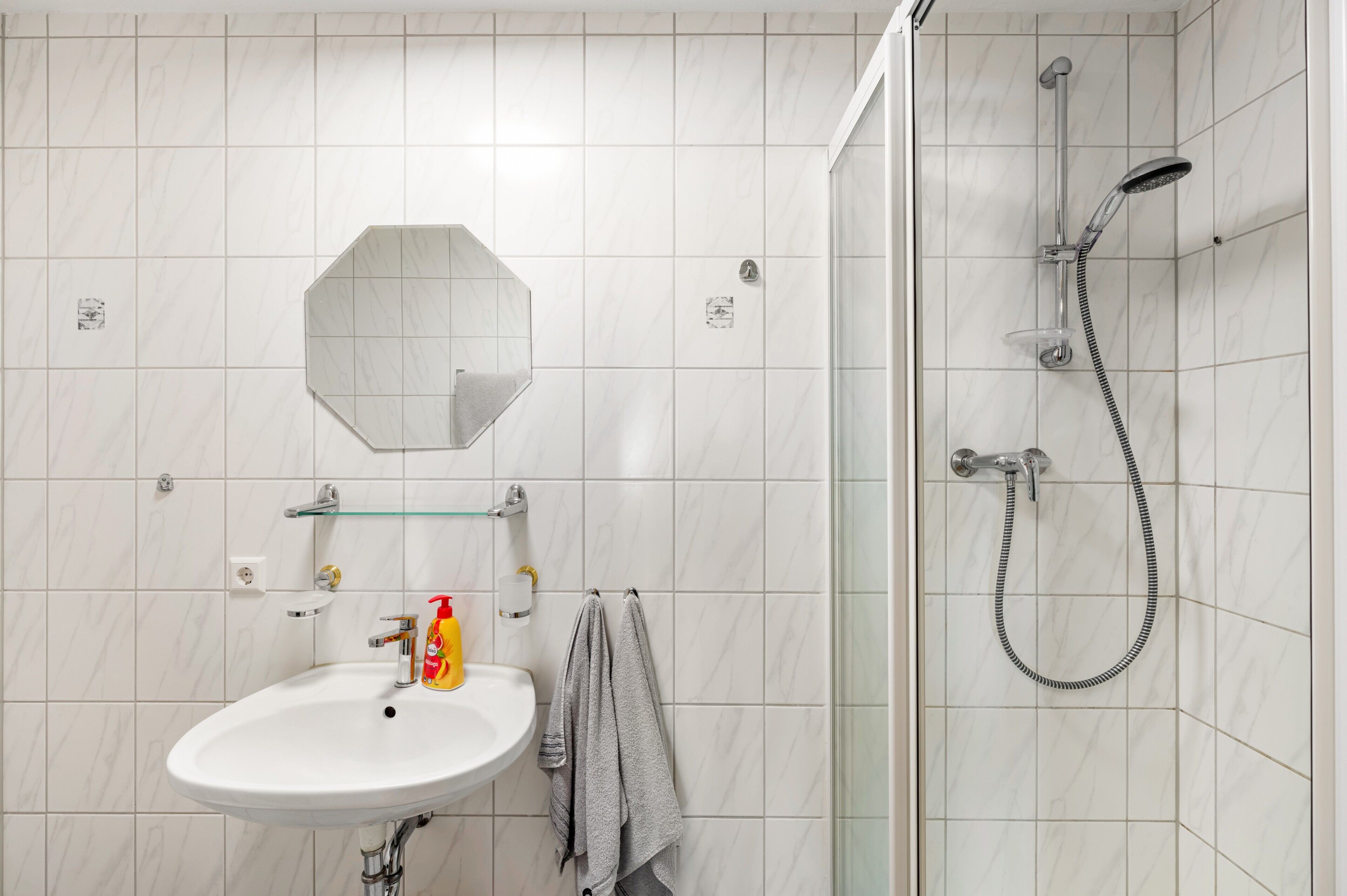 A bathroom with a white sink, a glass shelf, a towel rack, and a shower with a handheld showerhead.