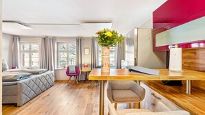 Une chambre d’hôtel moderne avec un lit, un bureau en bois, une chaise, un vase avec des fleurs et de grandes fenêtres.