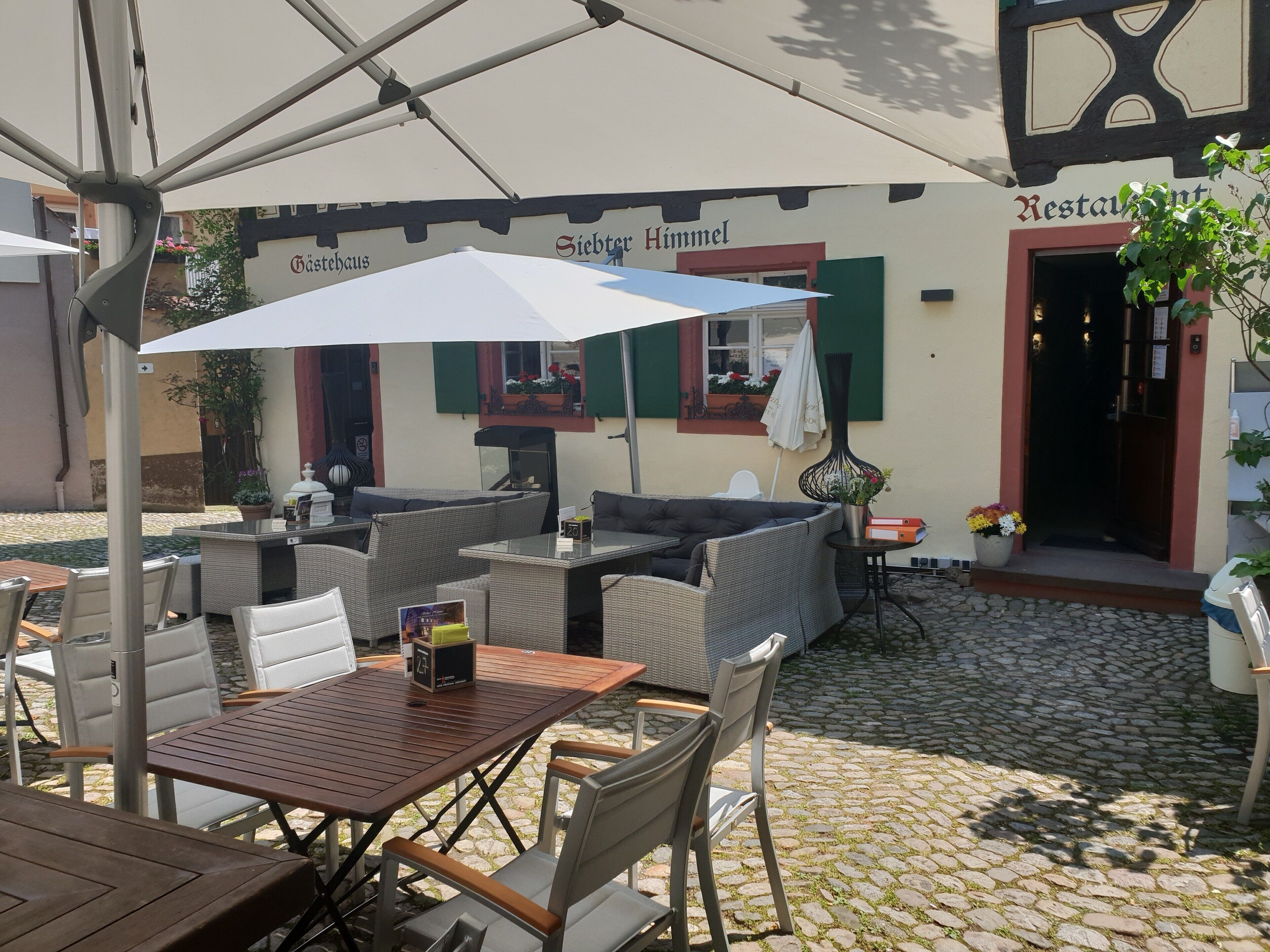 Ett utomhusområde med bord och stolar, ett vitt parasoll och en byggnad med en skylt som visar texten ”Siebter Himmel Restaurant”.