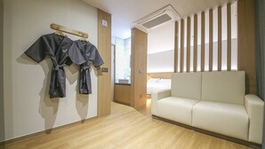 A hotel room with a wooden floor, a white sofa, and a rack with robes. - Brown Dot Sports Complex Branch Co., Ltd. (Jecheon)