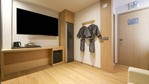 A hotel room with a flat-screen TV, a wooden desk, a small refrigerator, a coffee maker, and a robe hanging on a hook. - Brown Dot Sports Complex Branch Co., Ltd. (Jecheon)