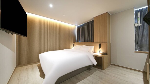 A modern hotel room with a large bed, wooden paneling on the walls, a flat-screen TV mounted on the wall, and a window with curtains. - Brown Dot Sports Complex Branch Co., Ltd. (Jecheon)