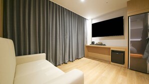 A modern hotel room with a flat-screen TV mounted on the wall, a wooden cabinet with a built-in refrigerator, a small kitchen area with a coffee maker, and a large window with grey curtains. - Brown Dot Sports Complex Branch Co., Ltd. (Jecheon)