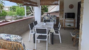 Un espacio cubierto para comer al aire libre con una mesa, sillas, un ventilador de techo y un televisor sobre un soporte de madera.