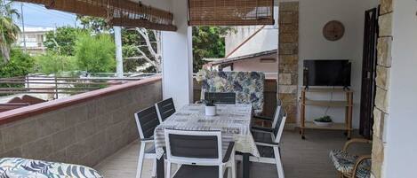 A covered outdoor dining area with a table and chairs, a ceiling fan, and a TV on a wooden stand.