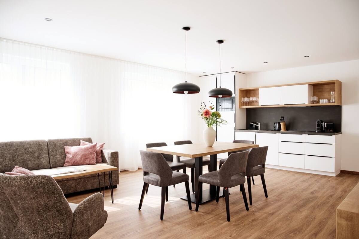 A modern living room with a sofa, dining table, and chairs. There is a kitchen area with cabinets and a microwave.