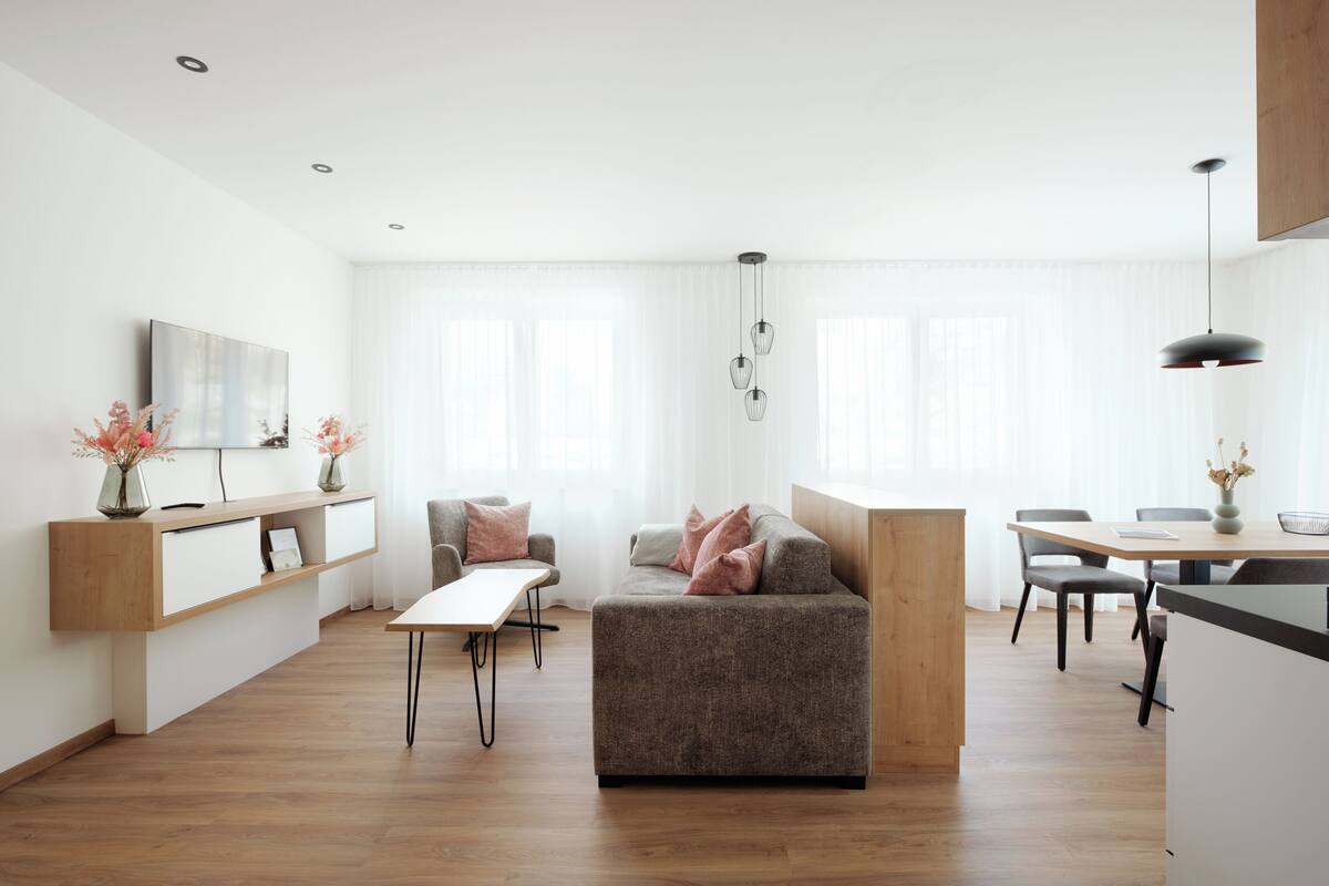A modern living room with a grey sofa, a wooden dining table, and a TV mounted on the wall.