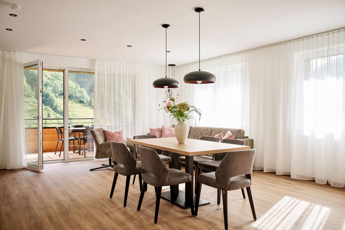 A dining area with a wooden table, grey chairs, and a vase with flowers. There is a sofa with cushions and a view of greenery outside.