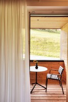Una terraza de madera con una mesa y una silla, una ventana que da a un campo cubierto de hierba, y una cortina medio bajada.