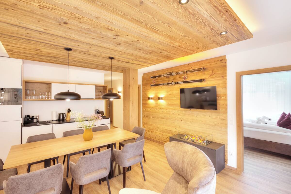 A modern kitchen with wooden ceiling, a dining area with a wooden table and chairs, a flat-screen TV mounted on the wall, and a cozy seating area with a sofa and a window with a view.