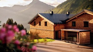 Une maison à deux étages, construite en briques, avec un toit en pente, un porche abrité et une vue sur les montagnes en arrière-plan.