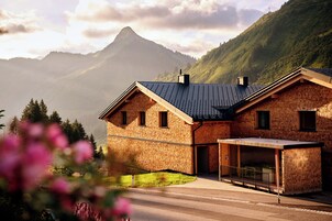 Una casa de ladrillo de dos pisos con techo inclinado, un porche cubierto y montañas al fondo.