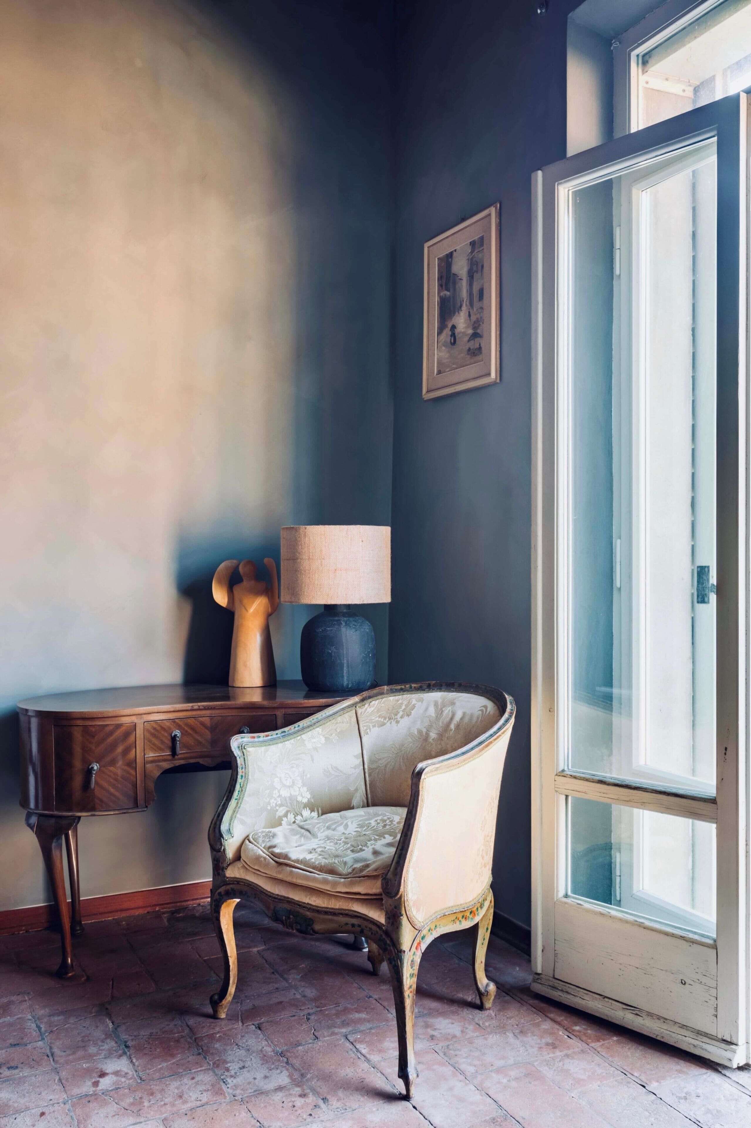 A corner room with a wooden desk, a cushioned chair, a lamp, and a door with a glass panel.