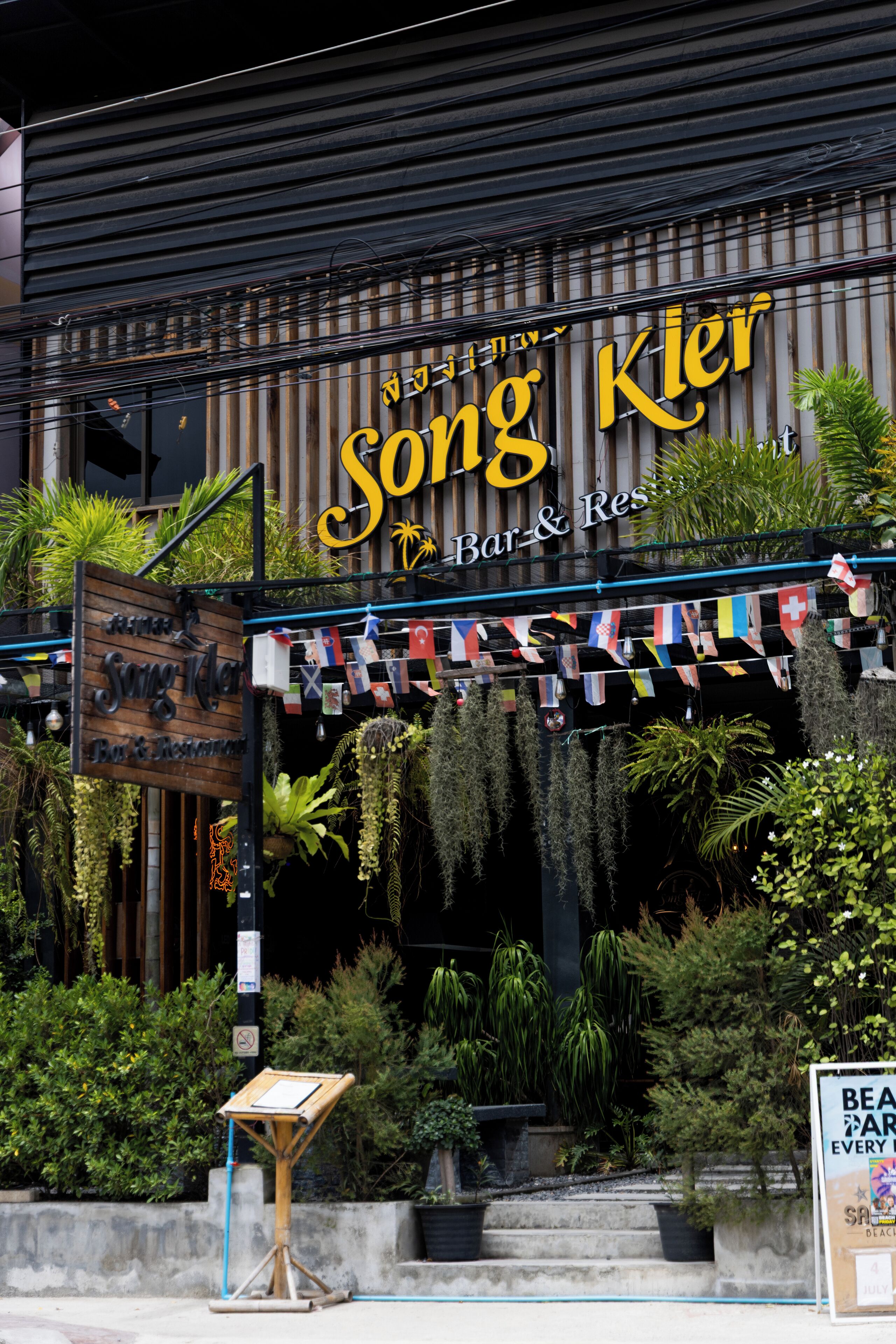 A restaurant named 'Song Kler Bar & Restaurant' with a signboard, outdoor seating, and a variety of plants.