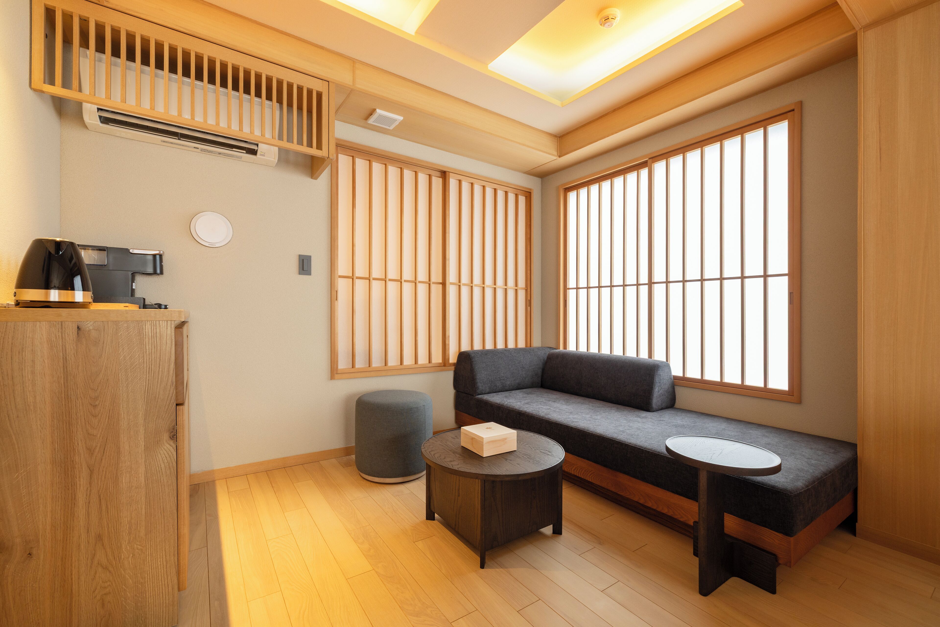 Une pièce moderne avec un plancher en bois, un canapé, une petite table ronde, un meuble et de grandes fenêtres équipées de stores verticaux.