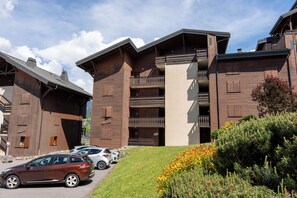 A multi-story building with balconies, a parking area with cars, and a grassy area with shrubs.