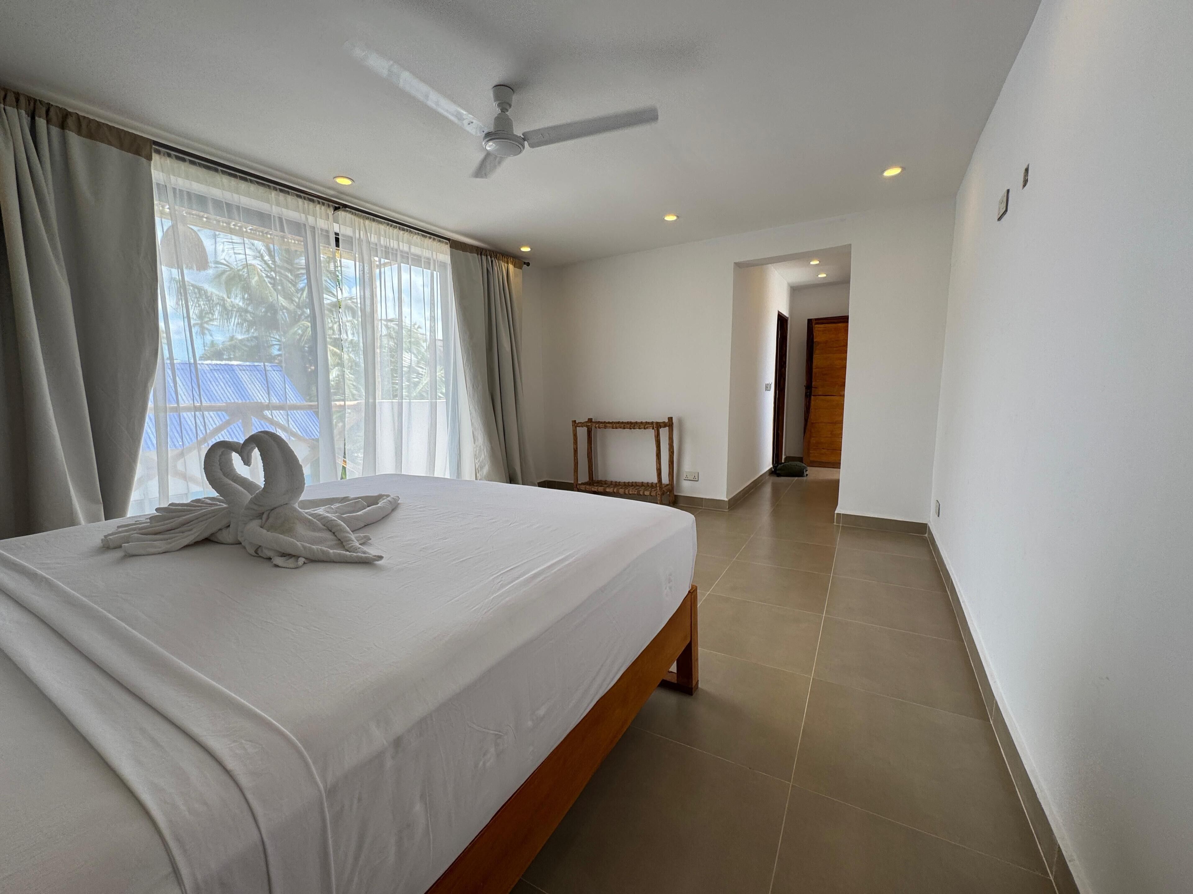 A hotel room with a bed, a ceiling fan, a view of the sea through the window, and a wooden chair.