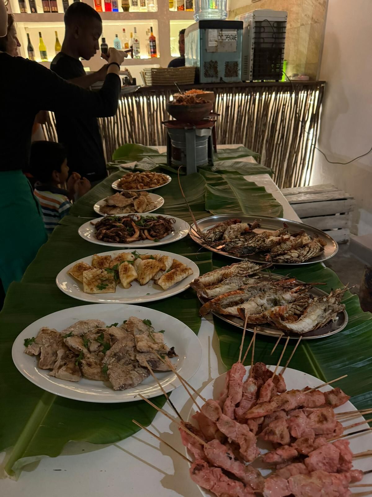 A buffet table with various dishes, including skewered meats and seafood, set on a table covered with banana leaves.