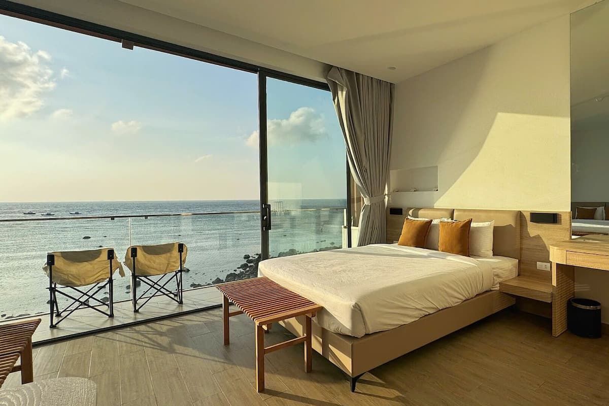 Ein modernes Hotelzimmer mit einem großen Bett, einem Holztisch, einem Sessel und einem Balkon mit Meerblick.